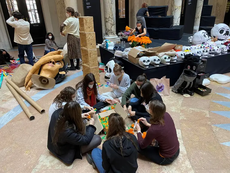 Eine Gruppe von mehreren Personen sitzt auf dem Boden und bastelt mit buntem Material. Um sie herum sind verschiedene Bastelmaterialien und dekorative Elemente verteilt. Im Hintergrund sind weitere Personen zu sehen, die ebenfalls an einem kreativen Projekt arbeiten. Die Atmosphäre ist aktiv und gemeinschaftlich.