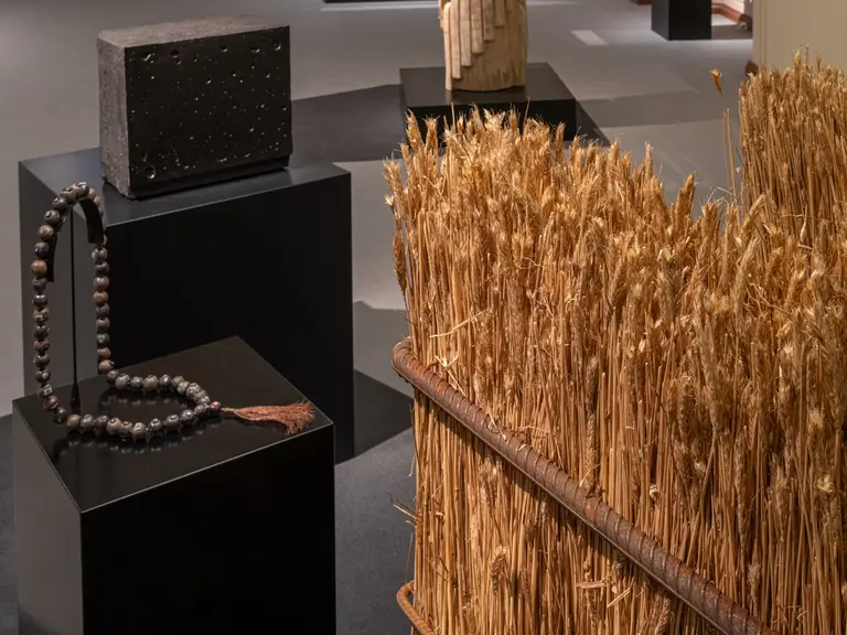 In einer Ausstellung sind verschiedene Kunstobjekte zu sehen. Auf einem schwarzen Sockel steht ein grauen Block, daneben eine dekorative Halskette aus Holzperlen. Im Hintergrund ist ein Arrangement aus getrocknetem Getreide zu sehen, das in einem großen Behälter angeordnet ist.