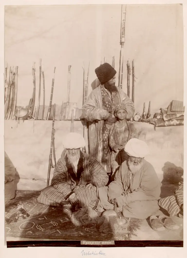 A historical photograph shows three men and a child inside a tent. One man, wearing a large turban and decorated robe, stands behind a seated child. Two elderly men sit on the floor, one with a beard, all dressed in traditional attire. The background features weapons and textiles.