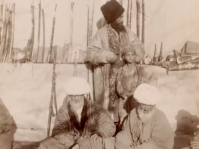 Die Schwarzweißaufnahme zeigt drei Männer in traditioneller Kleidung, darunter ein älterer Mann, der sitzt, und zwei weitere, die stehen. Im Hintergrund sind Gewehre und andere Gegenstände sichtbar. Die Szene vermittelt einen Eindruck von kulturellem Alltag in einer historischen Umgebung.