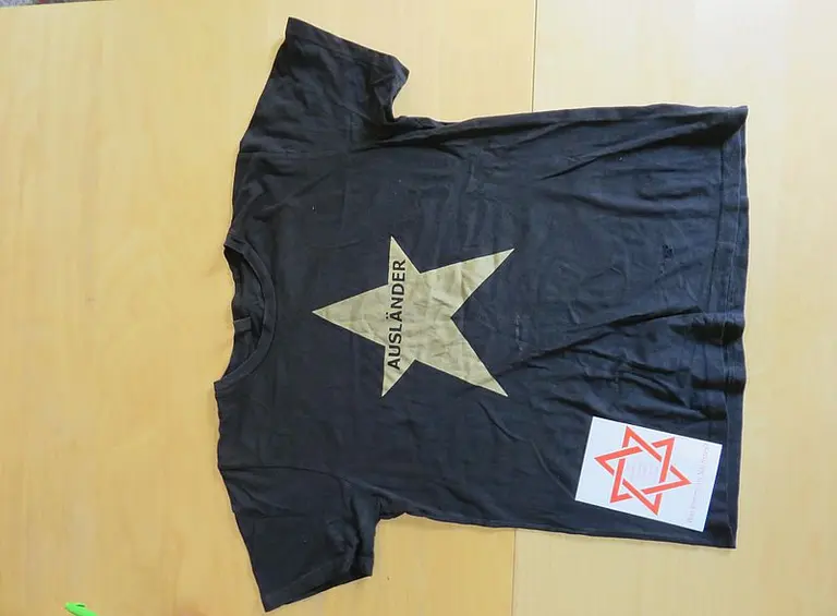 Ein schwarzes T-Shirt liegt flach auf einem Tisch. Es zeigt einen goldenen Stern mit dem Wort 