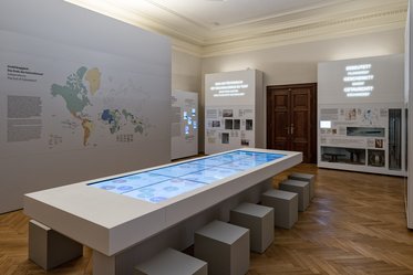 A modern exhibition space featuring a long interactive table with a touchscreen display. Surrounding walls showcase maps and informational panels. There are several small stools arranged around the table, and the atmosphere is bright and contemporary, highlighting educational content.