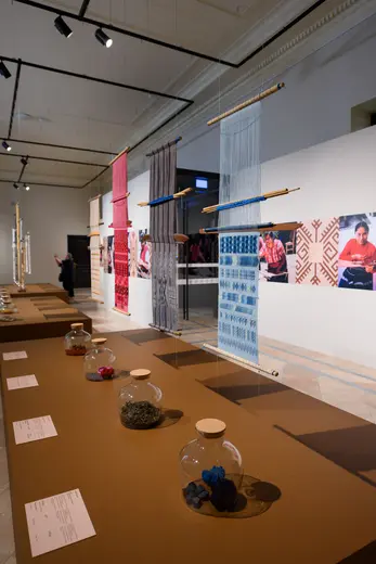 Eine Galerie mit gewebten Textilien in verschiedenen Farben hängt von Holzstangen. Im Vordergrund stehen Glaskaraffen mit Materialproben. Im Hintergrund sind Fotografien von Menschen, die das Handwerk ausüben. Die Ausstellung präsentiert traditionelle textile Kunst.