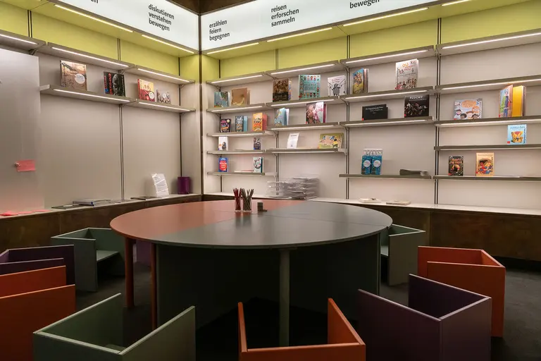 A cozy reading area featuring a round table surrounded by colorful chairs. Books are neatly arranged on shelves against the walls. The space is well-lit, creating an inviting atmosphere for reading and studying.