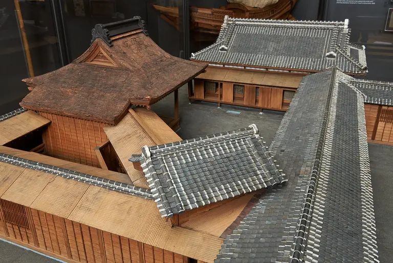 A detailed architectural model showcases traditional Japanese buildings, featuring distinct curved and tiled roofs, wooden structures, and intricate design elements. The model highlights the craftsmanship and layout of the buildings, emphasizing their historical and cultural significance.