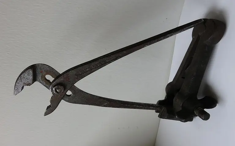 Das Bild zeigt eine rostige Zange mit einer speziellen Form. Die Zange hat lange Griffe und eine gebogene Vorderseite, die zum Greifen oder Halten von Gegenständen verwendet wird. Sie ist an einer Halterung befestigt, die an einer Oberfläche montiert ist.
