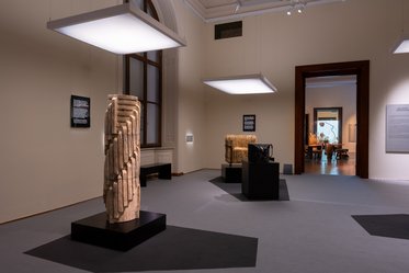 Modernes Kunstmuseum mit mehreren hölzernen Skulpturen auf dunklen Sockeln. Die Wände sind hell, und große Deckenlichter beleuchten die Ausstellungsstücke. Informationsbeschilderungen stehen neben den Skulpturen. Im Hintergrund sind weitere Räume des Museums sichtbar.