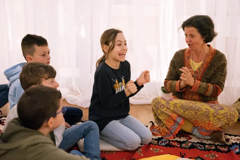 Eine Gruppe von Kindern sitzt auf dem Boden und hört aufmerksam zu. Ein Mädchen in einem schwarzen Shirt lächelt und gestikuliert fröhlich, während eine Frau in einem bunten, gestrickten Oberteil ihr zuschaut und mit beiden Händen klatscht.