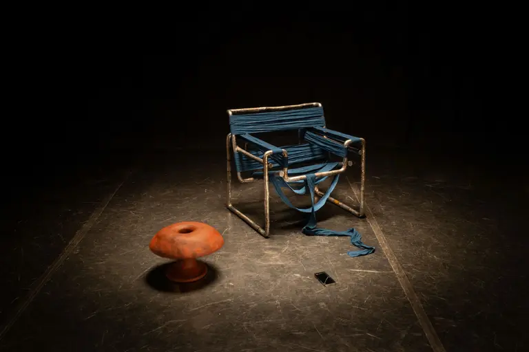 A blue-striped chair made of metal sits in a dimly lit space beside a round, orange stool. A blue ribbon drapes from the chair, and a black smartphone lies on the ground nearby. The setting has a minimalist, artistic feel.