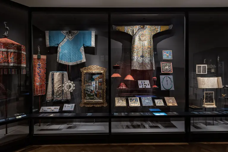 Das Bild zeigt eine Vitrine mit historischen Kleidungsstücken und Kunstwerken. Zu sehen sind farbenfrohe Roben, eine verzierte Rahmen und verschiedene Dokumente. Die Exponate sind sorgfältig angeordnet, um die kulturelle Bedeutung und die Handwerkskunst zu verdeutlichen.