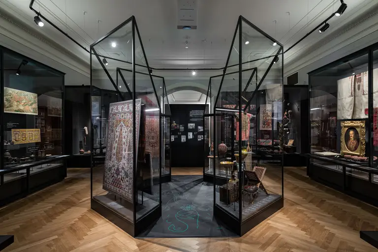 Eine Ansicht eines Ausstellungsraums mit mehreren Vitrinen, die historische Kunstwerke und Artefakte präsentieren. Die Vitrinen sind aus Glas und Metall, während der Boden mit Holzplanken verlegt ist. Im Hintergrund sind weitere kunstvolle Arbeiten zu sehen, die die Vielfalt der Ausstellung repräsentieren.