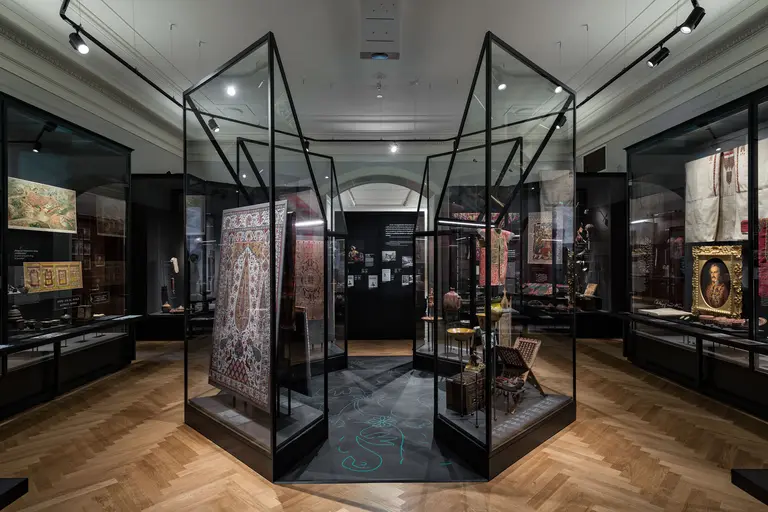 Eine Ansicht eines Ausstellungsraums mit mehreren Vitrinen, die historische Kunstwerke und Artefakte präsentieren. Die Vitrinen sind aus Glas und Metall, während der Boden mit Holzplanken verlegt ist. Im Hintergrund sind weitere kunstvolle Arbeiten zu sehen, die die Vielfalt der Ausstellung repräsentieren.