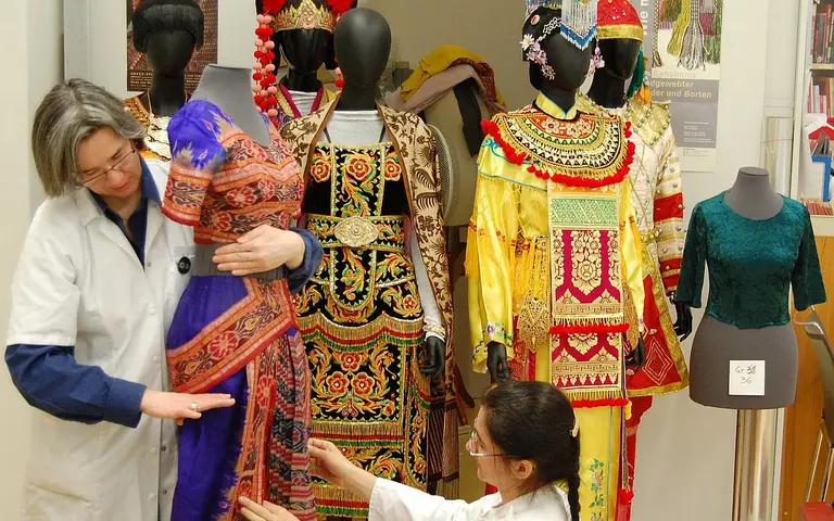 Two women in lab coats are examining and adjusting traditional garments displayed on mannequins. The colorful costumes, featuring intricate designs, represent various cultures. One woman holds a dress while the other inspects another outfit on a mannequin. The setting is an exhibition space.