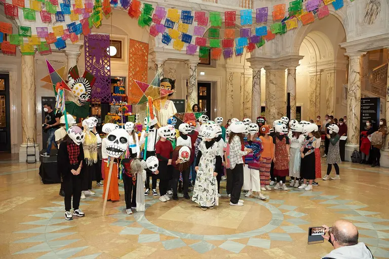 Eine Gruppe von Kindern und Erwachsenen in bunten Kostümen steht in einem festlich dekorierten Innenraum. Alle tragen große Totenkopf-Masken. Im Hintergrund hängen farbige Papierfähnchen, die eine festliche Atmosphäre schaffen. Es scheint eine kulturelle Feier zu sein.