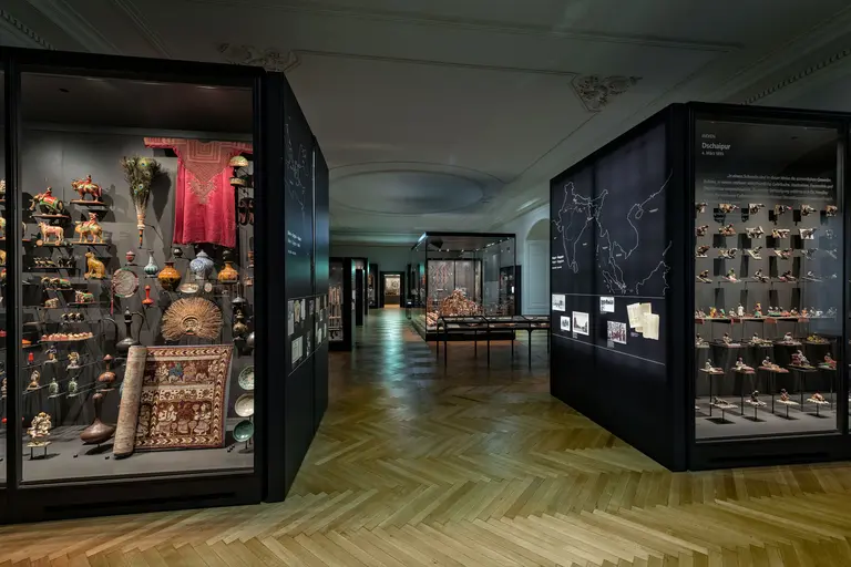 Das Bild zeigt einen Innenraum eines Museums mit mehreren Vitrinen, die vielfältige kulturelle Exponate präsentieren. Links sind farbenfrohe Kunstwerke und Textilien zu sehen, während rechts eine Sammlung von Objekten aus verschiedenen Materialien aufgereiht ist. Der Boden ist aus Holz und sorgt für eine warme Atmosphäre.