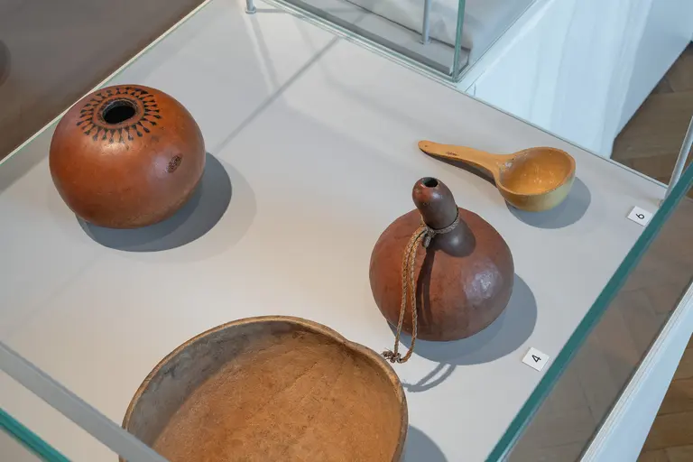 Ein Glasvitrinenboden mit mehreren traditionellen Gefäßen. Darunter sind zwei runde, bräunliche Schalen und ein kleinerer, eleganter Kegel. Eine Holzlöffel ist ebenfalls sichtbar. Der Hintergrund ist hell und deutlich, wodurch die Objekte in den Fokus gerückt werden.