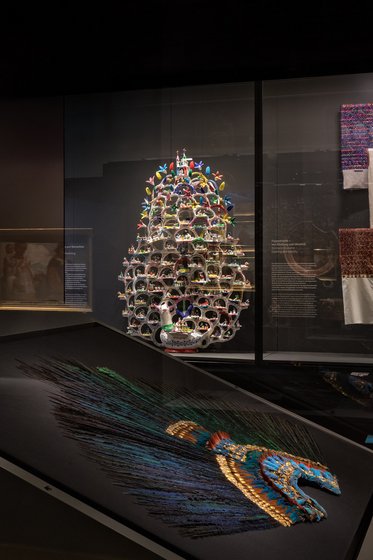 A colorful, intricate sculpture resembling a tower with floral and decorative elements sits in a glass display case. In the foreground, a dark, textured panel showcases vibrant threads in various colors, creating an abstract design. The overall setting suggests an art exhibit.