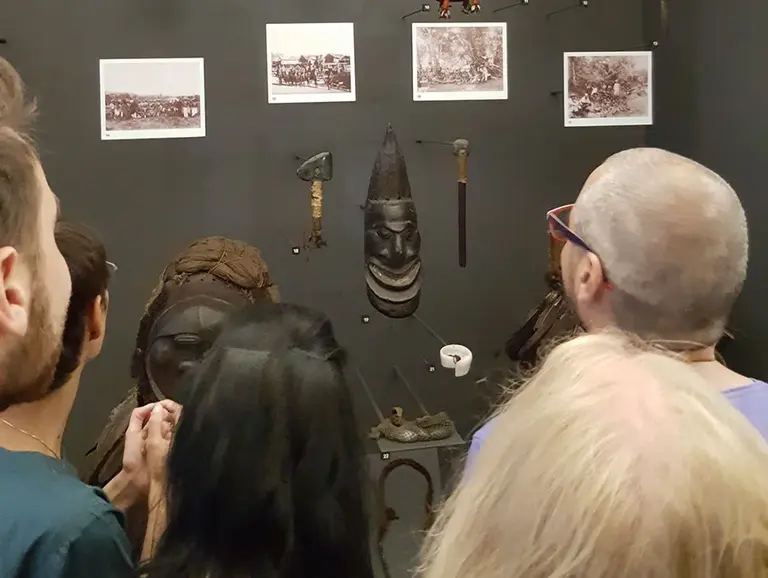 Die Abbildung zeigt eine Ausstellungswand mit verschiedenen traditionellen Werkzeugen und kulturanthropologischen Objekten. Darunter sind Fotografien, die vermutlich die historischen Verwendungen der Exponate zeigen. Mehrere Besucher schauen interessiert darauf.