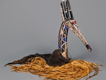 A decorative art piece resembling a bird's head, intricately crafted with patterned materials and colors. It features a long neck, a pointed beak, and tall, thin elements resembling horns. The base is made of natural fibers, adding texture and contrast to the design against a neutral background.