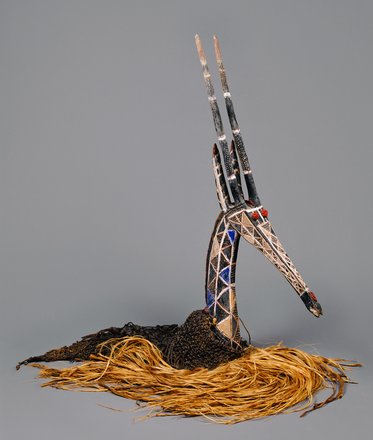 A decorative art piece resembling a bird's head, intricately crafted with patterned materials and colors. It features a long neck, a pointed beak, and tall, thin elements resembling horns. The base is made of natural fibers, adding texture and contrast to the design against a neutral background.