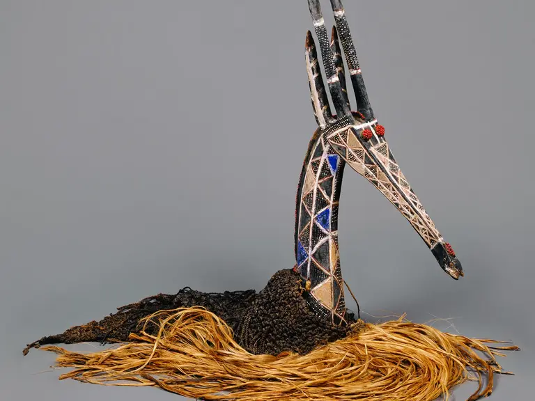 Das Bild zeigt eine künstlerische Skulptur eines Vogels mit einem langen Hals und einem auffälligen Kopf. Die Figur hat eine detaillierte Gestaltung mit bunten Mustern und ist auf einem Untergrund aus trockenen Pflanzenteilen platziert.