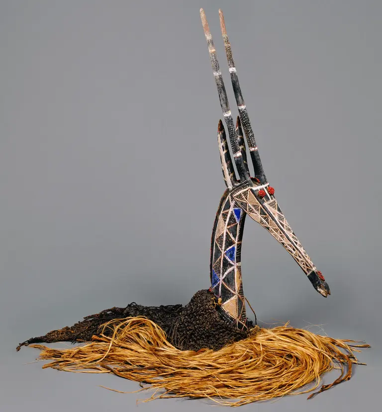 Das Bild zeigt eine künstlerische Skulptur eines Vogels mit einem langen Hals und einem auffälligen Kopf. Die Figur hat eine detaillierte Gestaltung mit bunten Mustern und ist auf einem Untergrund aus trockenen Pflanzenteilen platziert.