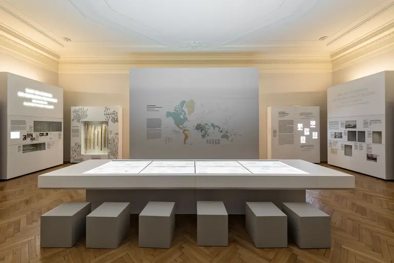 Das Bild zeigt eine moderne Ausstellung mit einem großen Tisch in der Mitte, umgeben von Informationswänden. Der Tisch ist mit interaktiven Displays ausgestattet. Im Hintergrund sind Karten und informative Texte sichtbar, die das Ausstellungsthema unterstützen. Der Boden ist aus Holz.