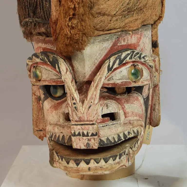 Eine traditionelle Maske mit einem detailliert geschnitzten Gesicht, großen Augen und einem offenen Mund. Oben trägt sie eine kunstvolle Kopfbedeckung aus geflochtenen Materialien und Textilien. Die Farbgestaltung umfasst Erdtöne und kräftige Farben.