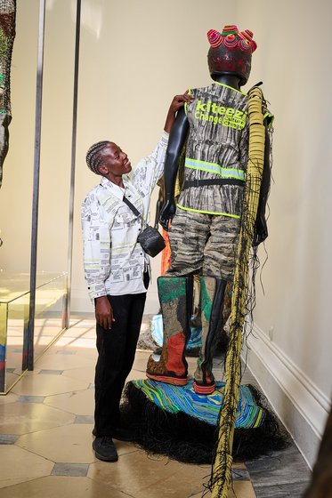 Ein Mann betrachtet eine Skulptur, die eine menschliche Figur darstellt. Die Figur ist in auffälligen, bunt gestalteten Kleidungsstücken mit verschiedenen Materialien bekleidet. Der Mann trägt ein gemustertes Hemd und hat eine Kamera um den Hals. Der Hintergrund ist neutral gehalten.
