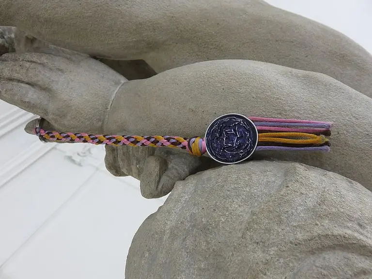 Eine geflochtene Armbänder mit bunten Fäden und einem runden, silbernen Anhänger liegt auf dem Arm einer Statue. Der Fokus liegt auf dem Armband, das eine lebendige, kreative Gestaltung zeigt. Die Statue selbst ist aus grauem Stein.