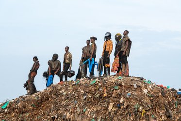 Gruppierung von Personen, die auf einem Müllhaufen stehen. Sie tragen unterschiedliche Kleidung und halten Gegenstände in ihren Händen. Der Himmel ist leicht bewölkt, und im Hintergrund sind Müllreste sichtbar. Der Blick vermittelt ein Gefühl von Dringlichkeit in Bezug auf Umweltfragen.