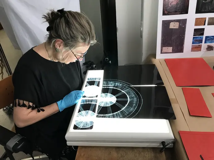 Eine Frau mit Brille und Handschuhen arbeitet konzentriert an einem Lichttisch, auf dem kreisförmige Zeichnungen oder Diagramme zu sehen sind. Um sie herum liegen rote und orangefarbene Blätter. Der Lichttisch dient zur Analyse oder Präsentation der Dokumente.