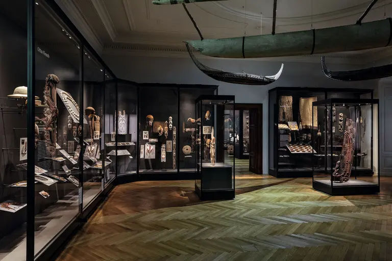 Ein Ausstellungsraum mit Glasvitrinen, die verschiedene kulturelle Artefakte und Kunstwerke zeigen. Im Hintergrund hängt ein traditionelles Boot. Der Boden ist aus Holz und verleiht dem Raum eine warme Atmosphäre.