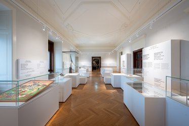 A spacious museum gallery featuring white display cases and informative panels. The floor is wooden, and the room is well-lit, highlighting various artifacts and exhibits arranged thoughtfully throughout the space. Natural light enters through large windows, creating an inviting atmosphere.