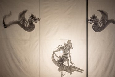 Silhouettes cast against a light backdrop show intricate shadow figures of a mythical creature on the left and a detailed character riding a crocodile in the center. The scene is framed by soft drapery, creating an artistic and mystical atmosphere.