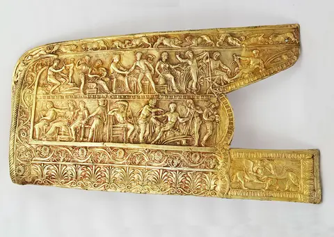A decorative gold relief featuring intricate scenes depicting figures engaged in various activities, including seated figures, animals, and decorative patterns. The piece showcases detailed artistry and craftsmanship typical of ancient Greek or Roman art. The overall shape is elongated with a rounded edge.