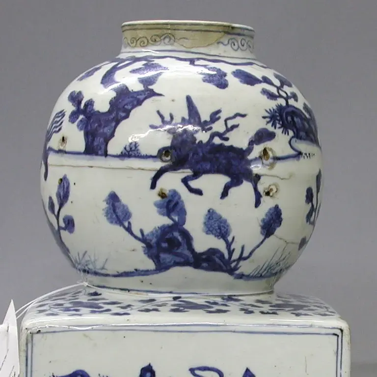 A two-tiered ceramic piece featuring blue and white patterns. The top is a rounded jar with floral and tree motifs, while the bottom is a square container depicting landscape scenes with figures and structures, all against a white background.