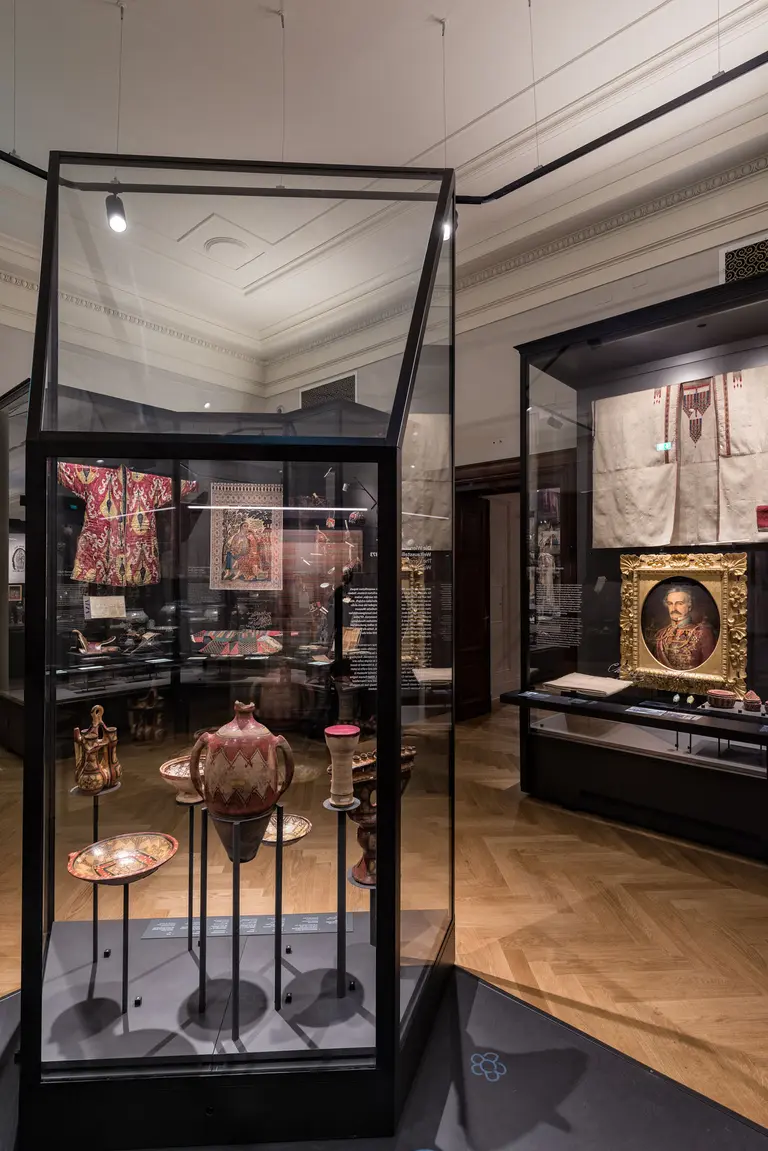 Die Abbildung zeigt eine Museumsausstellung mit Vitrinen. Im Vordergrund befindet sich eine gläserne Vitrine, die dekorierte Töpfe und Schalen zeigt. Im Hintergrund sind weitere Kunstwerke, darunter Gemälde und Textilien, sichtbar. Die Ausstellung hat einen historischen und kulturellen Charakter.