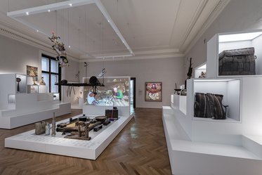 An art exhibition room featuring white display cases with various artifacts and items. A projection screen shows a scene with people engaging in activities. The space has wooden flooring and artwork on the walls, creating a bright and modern atmosphere.