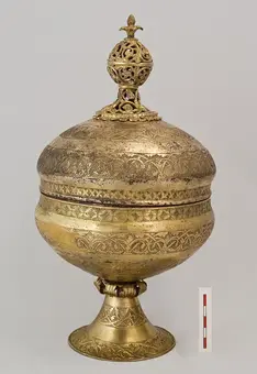 A decorative brass container with a round, bulbous body and a narrow neck. The lid features ornate detailing and a finial on top. The surface is intricately engraved with patterns, and it stands on a broad, flared base. A scale bar is included for size reference.