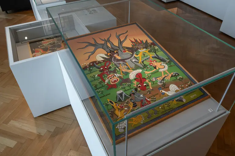 Ein Kunstwerk unter Glas, das eine lebendige Szene mit Tieren und Menschen zeigt. Die Komposition enthält farbenfrohe Figuren, darunter eine Mischung aus Mythologie und Natur, in einem abenteuerlichen Umfeld. Der Hintergrund ist grün, mit Bäumen und einem klaren Himmel.