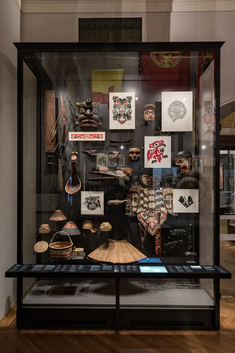 Eine Vitrine in einem Museum zeigt eine Sammlung traditioneller Kunstwerke, darunter Masken, Holzschnitzereien und ethnografische Objekte. Die Exponate stammen vermutlich aus indigenen Kulturen und sind kombiniert mit Zeichnungen und anderen kunstvollen Gegenständen.