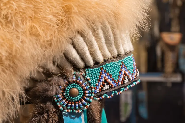 Das Bild zeigt einen Teil eines traditionell verzierten Kopfschmucks, der aus Federn und bunten Perlen besteht. Der Schmuck ist reich verziert mit türkisfarbenen und braunen Perlen, die geometrische Muster bilden. Der Hintergrund bleibt unscharf und lenkt nicht von dem Kopfschmuck ab.