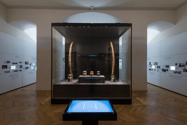 A well-lit museum exhibit featuring a glass display case with three masks and two large curved artifacts. Surrounding walls have historical photographs and information panels. A digital touchscreen display is positioned in front of the case, showcasing interactive content. The floor is wooden, enhancing the elegant atmosphere.