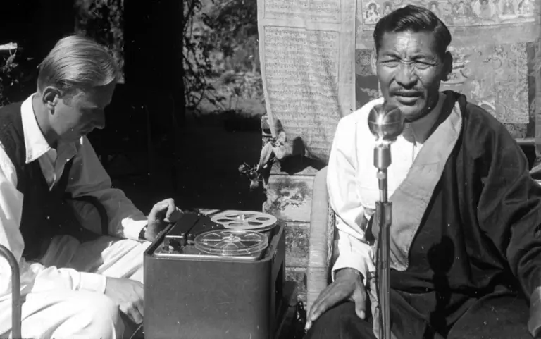 Ein Mann im traditionellen Gewand sitzt vor einem Mikrofon, während ein anderer Mann sich an einem Tonbandgerät beschäftigt. Die Szene vermittelt eine Verbindung zwischen Mundart und moderner Technik.