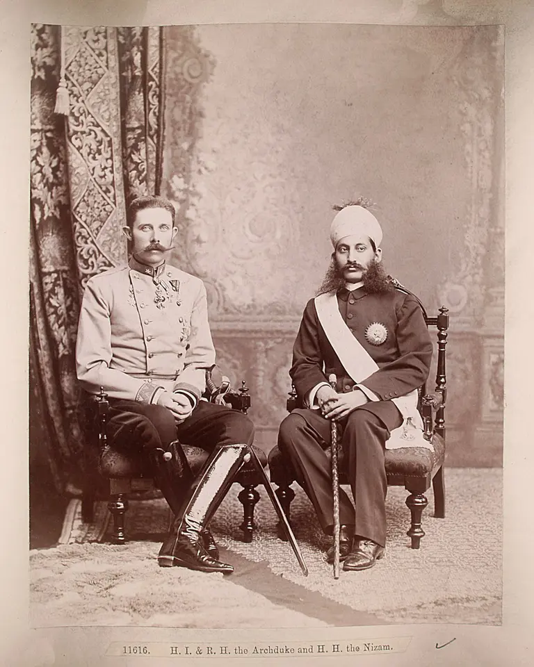 Historisches Foto, das zwei Männer in formellen Kleidungen zeigt, die in einer eleganten Umgebung sitzen. Der Mann links trägt eine militärische Uniform, der Mann rechts einen traditionellen festlichen Anzug mit einem Kopfschmuck. Beide schauen ernst in die Kamera.