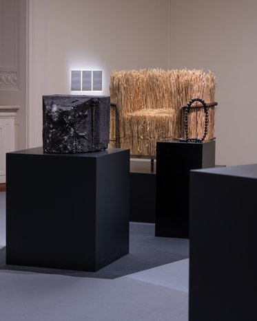In einem Ausstellungsraum stehen mehrere Skulpturen auf schwarzen Podesten. Links ein schwarzes, blockartiges Objekt, in der Mitte ein Stuhl aus Stroh mit dunklen Griffen. Die Umgebung ist schlicht und modern gestaltet.