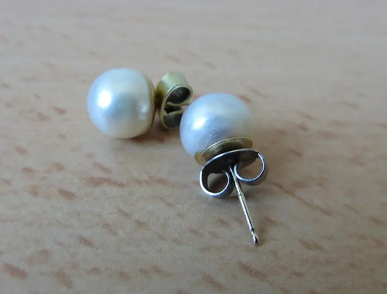 Zwei elegante Perlenohrringe liegen auf einer Holzoberfläche. Die Perlen sind rund und haben einen schimmernden, weißen Farbton. Die Bleistifte sind mit einer goldenen Fassung verbunden, die den Ohrringstift hält.