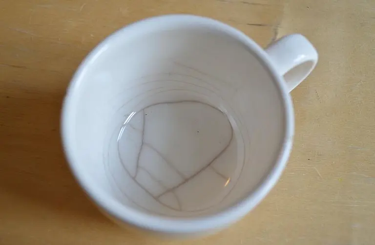 Eine weiße Tasse von oben betrachtet, die fast leer ist. Im Inneren sind feine Risse sichtbar, die das Design der Tasse bilden. Die Tasse steht auf einem Holzuntergrund.