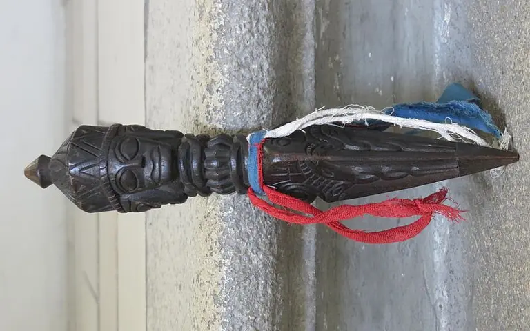Eine dunkle Holzfigur in Form eines stilisierten menschlichen Gesichts, verziert mit einer spitzen Oberseite. Um die Figur sind mehrere Farbbanden in Rot und Blau gebunden, die ihr zusätzlichen Kontrast und Textur verleihen. Die Figur steht auf einem grauen Untergrund.